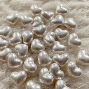 May include: A collection of white pearl heart-shaped beads on a white fluffy surface.