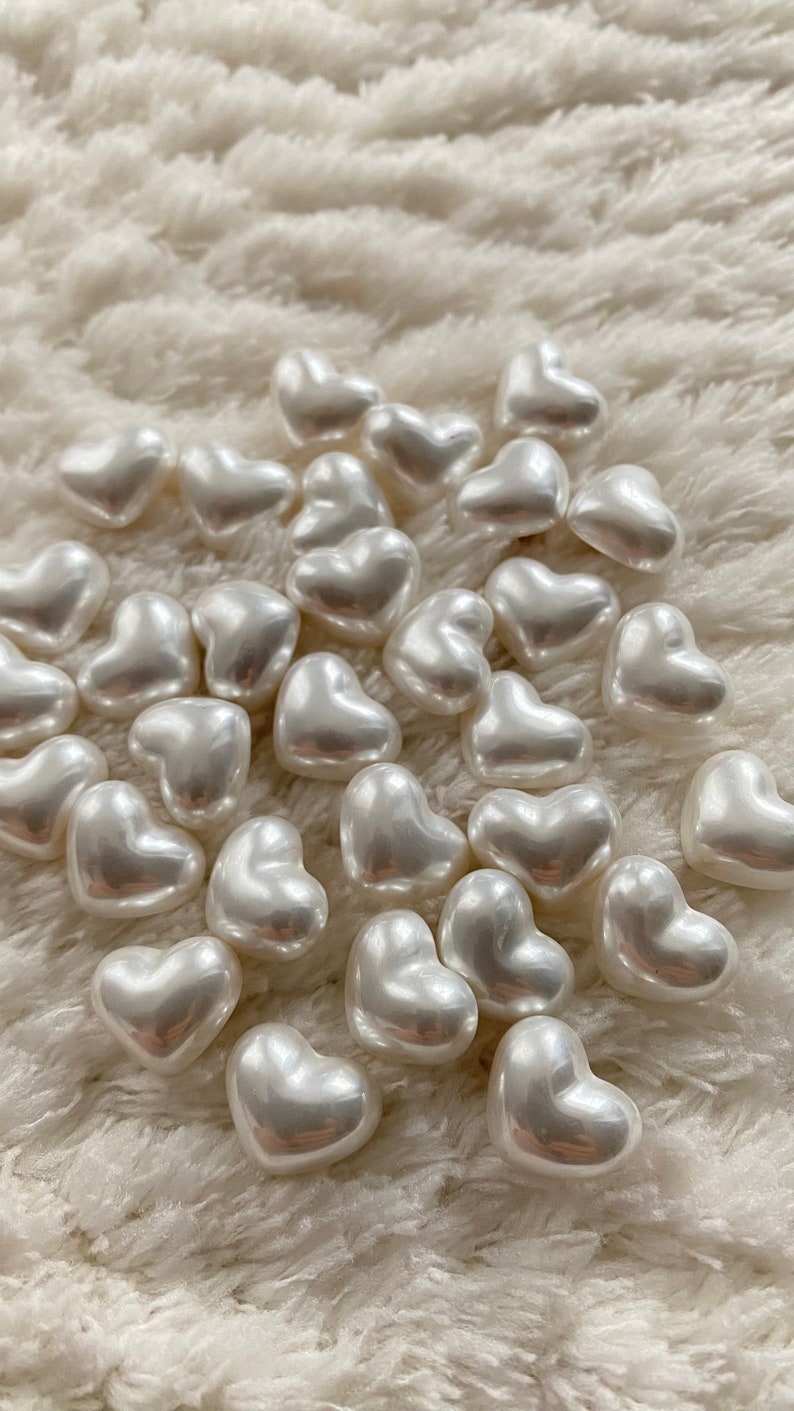 May include: A collection of white pearl heart-shaped beads on a white fluffy surface.
