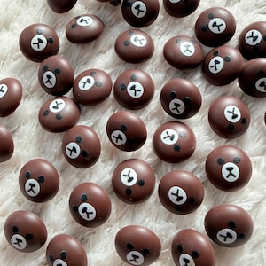 May include: Brown, round buttons featuring a bear face design. Each button has a white face with black eyes and a black nose. The buttons are scattered on a white, fluffy surface.