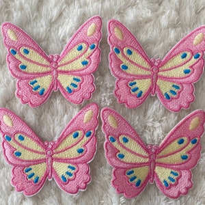 May include: Four pink and yellow embroidered butterfly patches with blue accents. The patches are on a white fluffy surface.