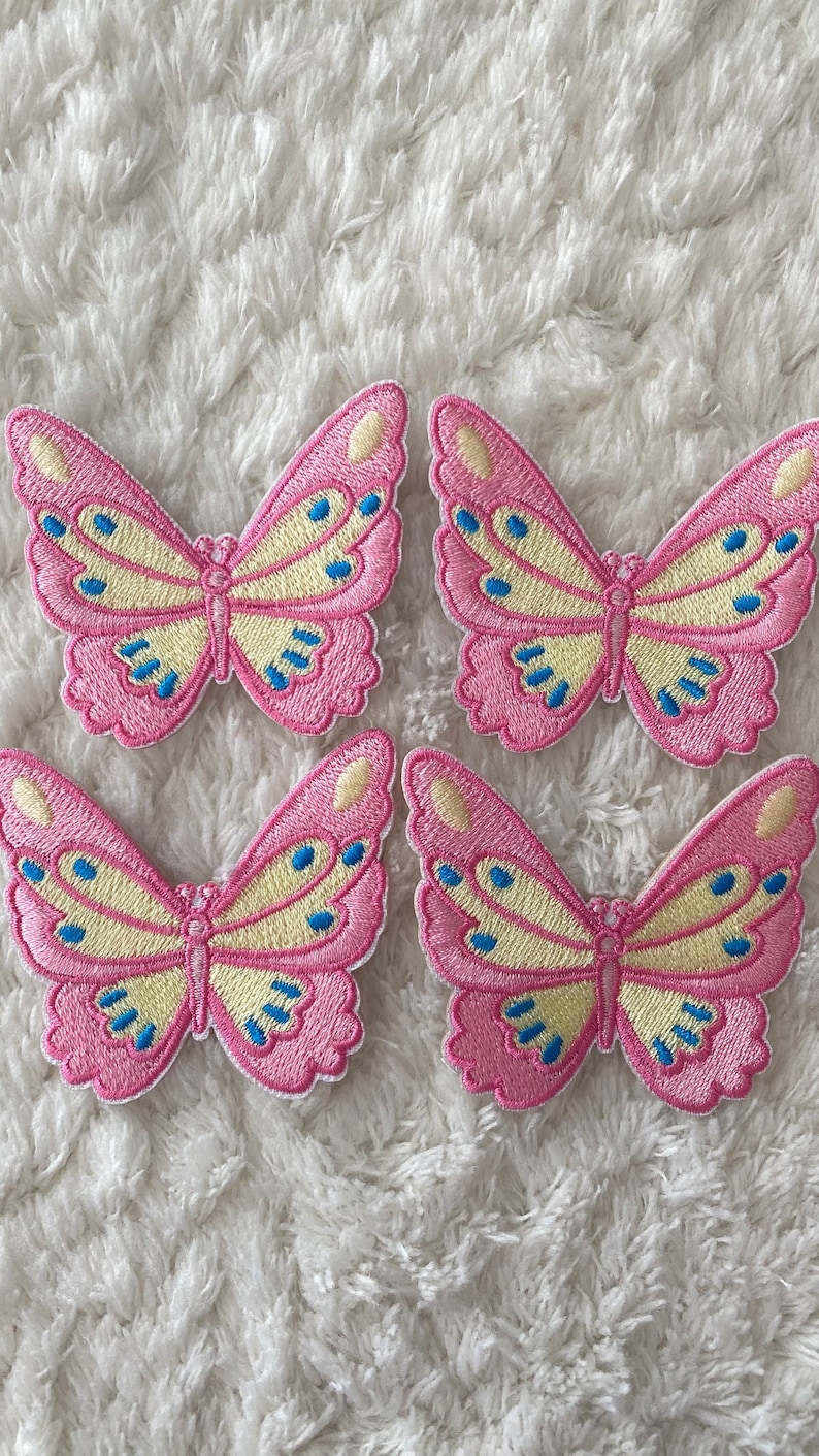May include: Four pink and yellow embroidered butterfly patches with blue accents. The patches are on a white fluffy surface.