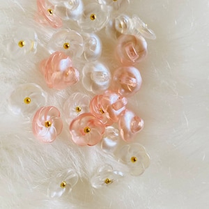Könnte beinhalten: Eine Sammlung von rosa und durchsichtigen, blumenförmigen Knöpfen mit goldenen Mittelpunkten. Die Knöpfe sind auf einer weißen, flauschigen Oberfläche verstreut.