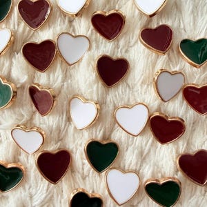 May include: A collection of heart-shaped embellishments in white, burgundy, and green, each with a gold-colored border. The hearts are scattered on a textured, off-white surface, creating a visually appealing pattern.
