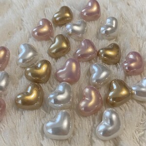 May include: A collection of heart-shaped pearl beads in shades of pink, white, and gold. The beads are arranged on a white fuzzy surface.