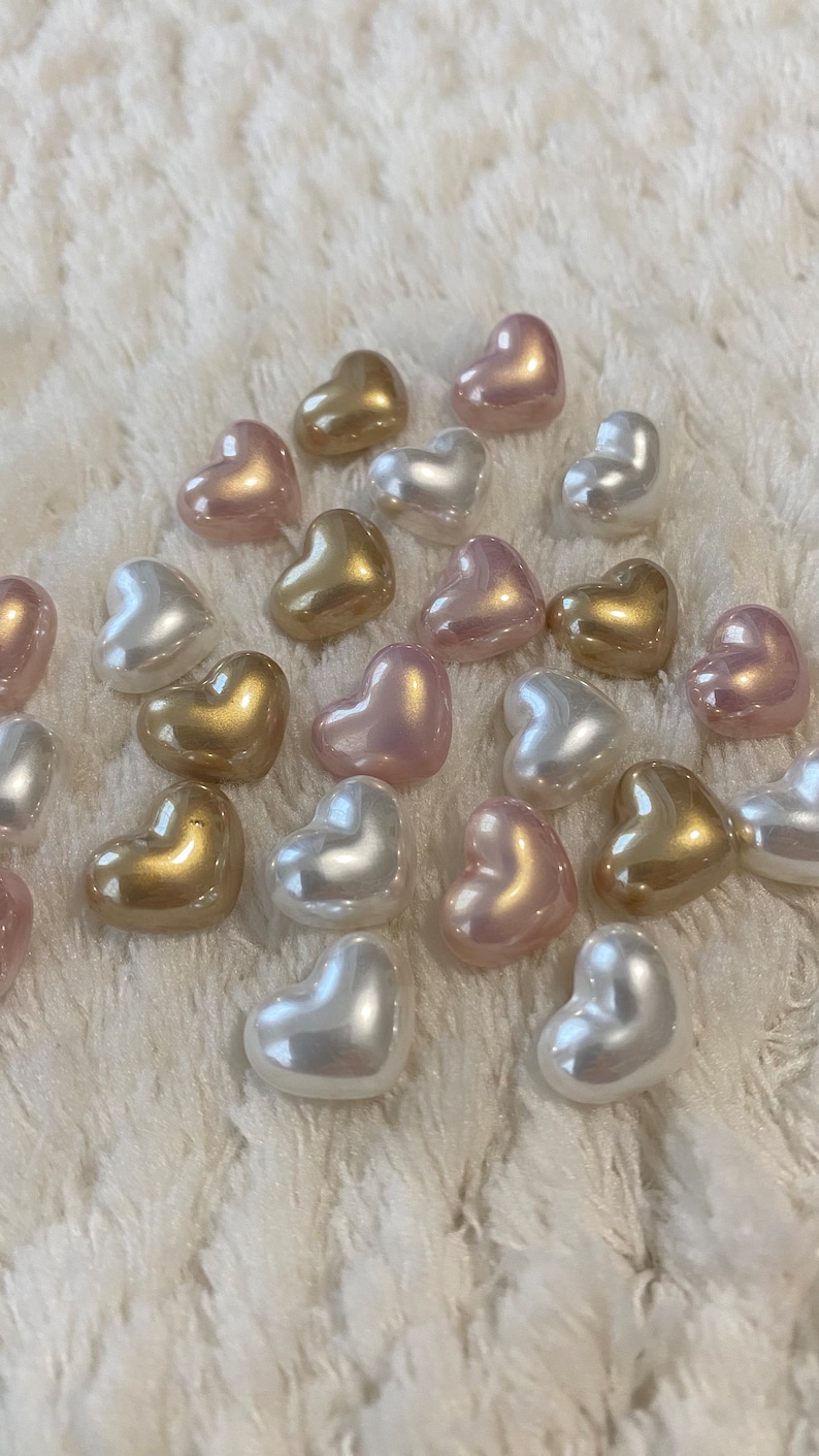 May include: A collection of heart-shaped pearl beads in shades of pink, white, and gold. The beads are arranged on a white fuzzy surface.