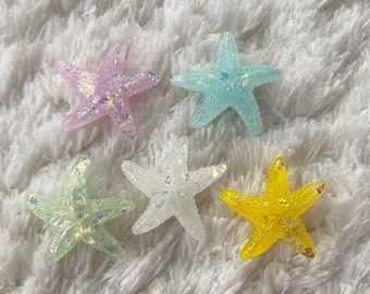 May include: Five sparkly starfish in different colors: pink, light blue, white, green, and yellow. The starfish are on a white fluffy surface.