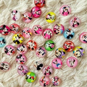 Op de afbeelding: Een verzameling kleurrijke, ronde kralen met afbeeldingen van Mickey en Minnie Mouse. De kralen zijn in verschillende kleuren, waaronder roze, geel, rood en blauw, en zijn verspreid over een witte, zachte ondergrond. Ideaal voor knutselen en het maken van sieraden.