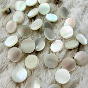 May include: A collection of iridescent, round mother-of-pearl buttons scattered on a white, fluffy surface. The buttons vary in size and have a lustrous, pearly sheen, reflecting shades of white, cream, and subtle pastel hues. Some buttons have metal shanks.