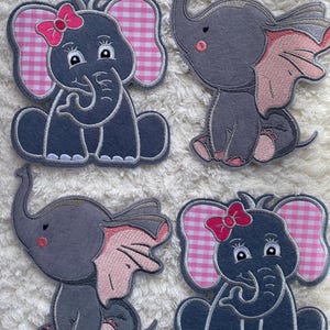 Può includere: Quattro applicazioni di feltro grigio a forma di elefante con orecchie rosa e fiocchi rosa a quadri. Due degli elefanti hanno fiocchi rosa a quadri sulla testa.