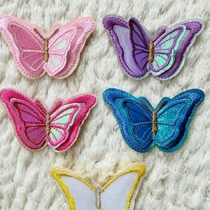 May include: Five embroidered butterfly patches in various colors: pink, purple, blue, and white with gold accents. The patches have a textured appearance and are arranged on a white, fluffy surface. These patches are ideal for adding a decorative touch to clothing or accessories.