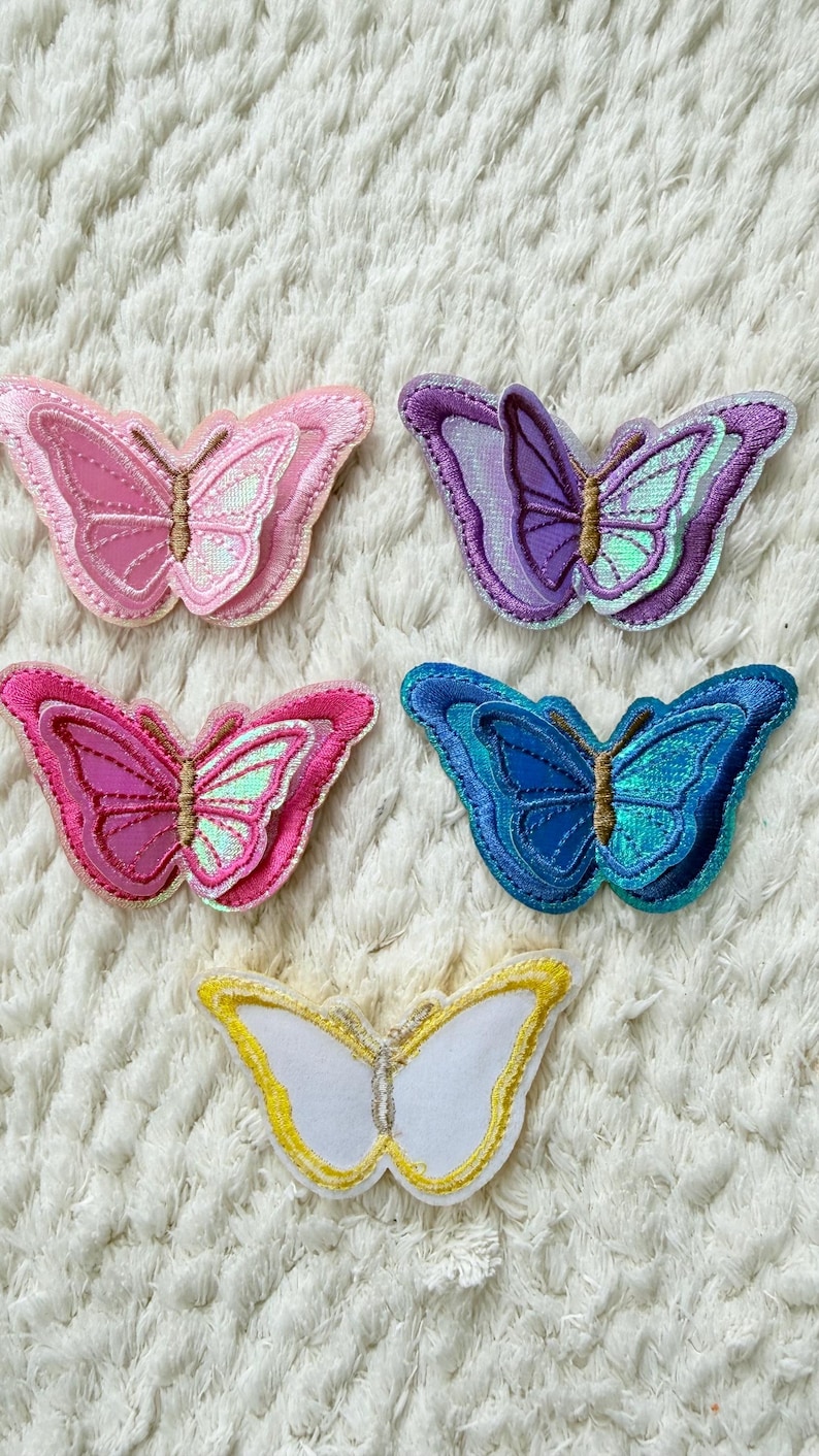 May include: Five embroidered butterfly patches in various colors: pink, purple, blue, and white with gold accents. The patches have a textured appearance and are arranged on a white, fluffy surface. These patches are ideal for adding a decorative touch to clothing or accessories.