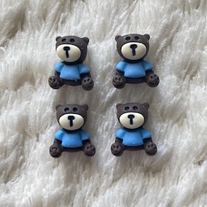 Op de afbeelding: Vier bruine teddybeer-knoppen met blauwe shirts. De knoppen liggen op een wit, pluche oppervlak.