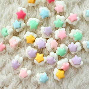Könnte beinhalten: Eine Ansammlung farbenfroher sternförmiger Verzierungen. Jeder Stern hat eine andere Pastellfarbe, darunter Rosa, Gelb, Blau, Grün und Lila, und ist auf einer weißen wolkenförmigen Basis angeordnet. Der Hintergrund ist eine weiche, weiße, strukturierte Oberfläche.