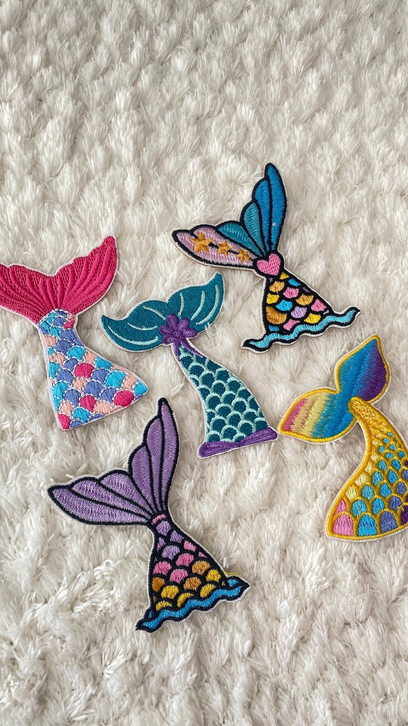 May include: Five colorful embroidered mermaid tail patches. The tails are in various colors, including pink, blue, green, purple, and yellow. Each tail has a different design and pattern.
