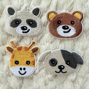 Puede incluir: Cuatro parches bordados de animales: un mapache, un oso, una jirafa y un perro. El mapache es gris y negro, el oso es marrón, la jirafa es amarilla y naranja, y el perro es marrón y negro. Cada parche tiene un borde blanco.