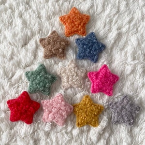 May include: Ten fluffy star-shaped ornaments in various colors, including red, pink, orange, yellow, green, blue, and gray. The stars are arranged in a circle on a white fluffy surface.