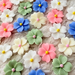 Puede incluir: Una colección de pequeños adornos florales coloridos en varios tonos de rosa, blanco, azul, verde y amarillo. Cada flor tiene un centro amarillo y un centro rosa o verde. Las flores están dispuestas sobre una superficie suave y blanca texturizada.