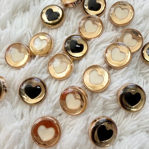 May include: A collection of round, decorative buttons with a gold-colored rim. Each button features a heart-shaped design in either black or white. The buttons are scattered on a white, fluffy surface.