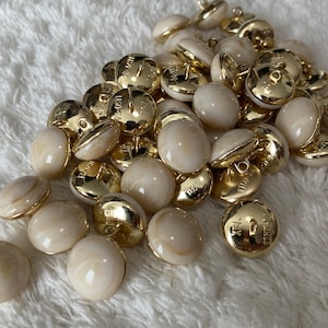 Puede incluir: Un montón de botones blancos y dorados con un acabado nacarado. Los botones tienen un borde dorado y un pequeño bucle en la parte superior.