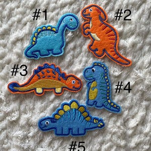 May include: Five felt dinosaur patches in various colors and designs. The patches are numbered 1 through 5. The dinosaurs are blue, orange, and green.