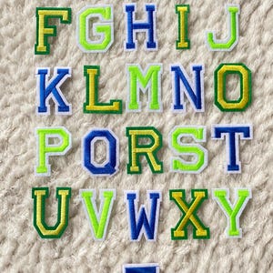 Può includere: Un set di 26 lettere dell'alfabeto in feltro colorato. Le lettere hanno un contorno bianco con un riempimento verde, giallo o blu. Le lettere sono disposte in ordine alfabetico.