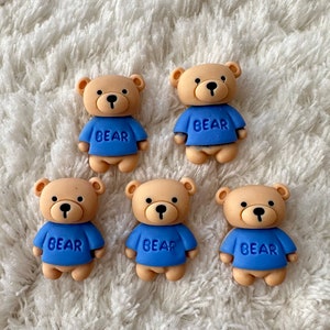 Puede incluir: Cinco figuritas de ositos de peluche marrones que llevan camisas azules con la palabra "BEAR" impresa. Los osos están dispuestos en semicírculo sobre una superficie blanca y esponjosa.