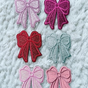 May include: Six embroidered bow patches in various shades of pink, red, and silver. The bows have a glittery texture and are arranged on a white, textured surface. These patches are suitable for clothing or craft projects.