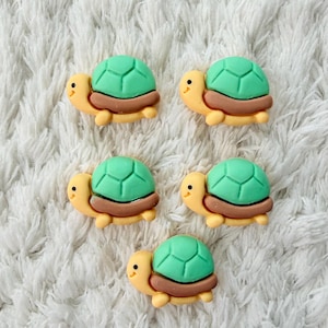 Könnte beinhalten: Fünf kleine Schildkröten-Verzierungen. Jede Schildkröte hat einen grünen Panzer, einen beigen Körper und eine braune Basis. Die Schildkröten haben kleine schwarze Augen und sind auf einer weißen, strukturierten Oberfläche angeordnet.