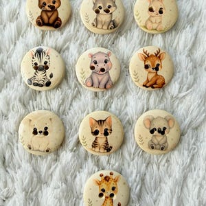 May include: Ten cream-coloured, round buttons, each with a different cute animal illustration. Animals include a bear, wolf, rabbit, zebra, hippo, deer, polar bear, cat, koala, and giraffe. The buttons are arranged on a white, textured surface.