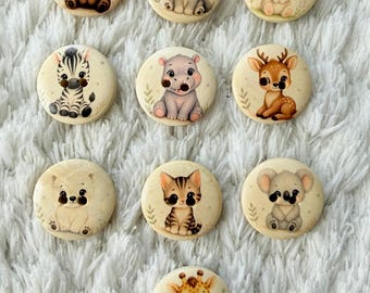 May include: A collection of ten round, cream-colored buttons, each featuring a different adorable animal illustration. Animals include a bear, wolf, rabbit, zebra, hippo, deer, polar bear, cat, koala, and giraffe. The buttons are arranged on a white, textured surface.