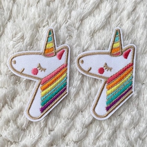 Può includere: Due toppe ricamate di unicorno bianche con criniere arcobaleno. Gli unicorni hanno gli occhi chiusi e le guance rosa. Le toppe sono su una superficie bianca e pelosa.