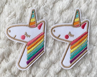 Può includere: Due toppe ricamate di unicorno bianche con criniere arcobaleno. Gli unicorni hanno gli occhi chiusi e le guance rosa. Le toppe sono su una superficie bianca e pelosa.