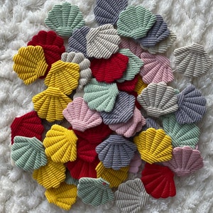 Könnte beinhalten: Eine Sammlung gehäkelter Muscheln in verschiedenen Farben, darunter Gelb, Rot, Grün, Rosa und Grau. Die Muscheln sind auf einer weißen, flauschigen Oberfläche zu einem Haufen zusammengelegt.