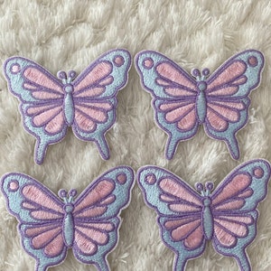 May include: Four pastel purple and light blue butterfly iron-on patches. The butterflies have pink accents and are arranged in a square pattern.
