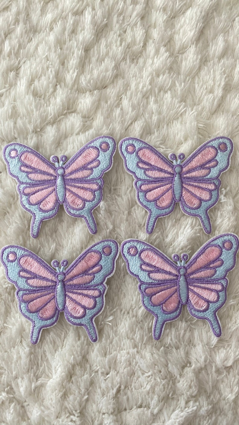 May include: Four pastel purple and light blue butterfly iron-on patches. The butterflies have pink accents and are arranged in a square pattern.