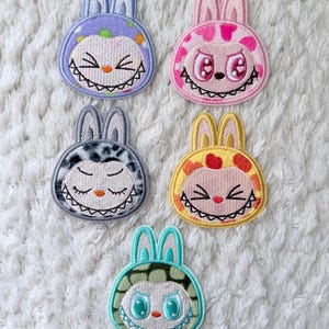 Könnte beinhalten: Fünf bunte Hasenaufnäher mit gestickten Details. Jeder Aufnäher zeigt ein einzigartiges Design mit einem lächelnden Gesicht, darunter ein lila, rosa, grauer, gelber und türkisfarbener Hase. Die Aufnäher haben eine skurrile und verspielte Ästhetik.