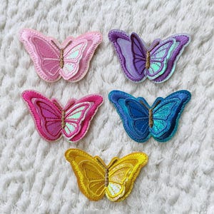 May include: Five embroidered butterfly patches in shades of pink, purple, blue, and yellow. Each butterfly has detailed wing patterns and a shimmering, iridescent center. The patches are arranged on a white, textured surface.