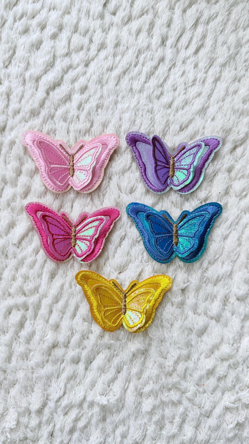 May include: Five embroidered butterfly patches in shades of pink, purple, blue, and yellow. Each butterfly has detailed wing patterns and a shimmering, iridescent center. The patches are arranged on a white, textured surface.