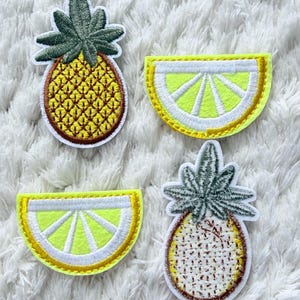 May include: Four embroidered fruit patches: two pineapples and two lemon slices. The pineapples have green tops and yellow bodies, while the lemon slices have yellow centres and white and yellow borders. Set against a white, textured backdrop.