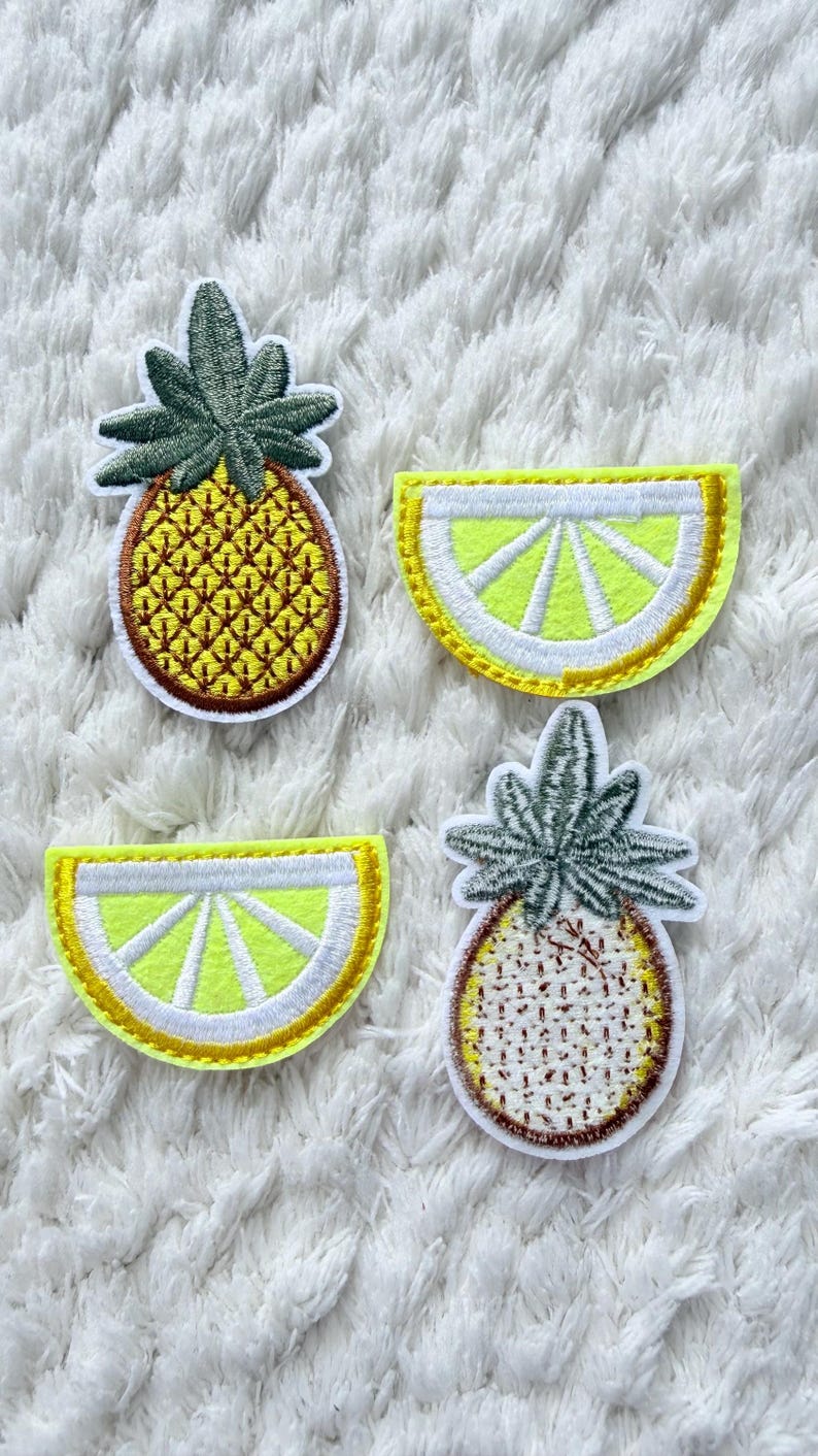 May include: Four embroidered fruit patches: two pineapples and two lemon slices. The pineapples have green tops and yellow bodies, while the lemon slices have yellow centres and white and yellow borders. Set against a white, textured backdrop.