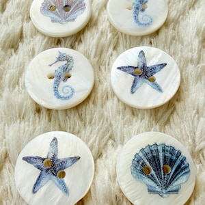 Peut inclure: Six boutons ronds blancs à la finition nacrée, chacun doté de deux trous. Les boutons sont ornés d'illustrations bleues de vie marine, notamment des hippocampes, des étoiles de mer et des coquillages, disposés en trois rangées de deux.