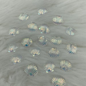 Puede incluir: Una colección de adornos iridiscentes en forma de concha de mar sobre una superficie blanca y esponjosa. Las conchas son de varios tamaños y tienen un acabado nacarado.