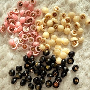Può includere: Una collezione di bottoni assortiti in rosa, crema e nero. I bottoni hanno una forma rotonda con gambo dorato. I bottoni rosa hanno un effetto marmorizzato, mentre i bottoni neri hanno un design maculato.