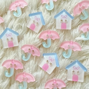 Può includere: Una collezione di bottoni piatti a due fori a forma di casette azzurre e rosa e ombrelli rosa con manici azzurri. Ogni bottone presenta un piccolo dettaglio a fiocco rosa. I bottoni a forma di casa hanno una porta rosa, quelli a ombrello un manico curvo.