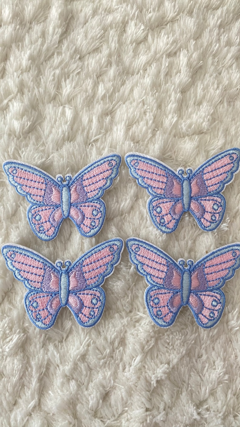 May include: Four pink and blue butterfly iron-on patches. The butterflies have a light pink and purple striped pattern with a blue outline.