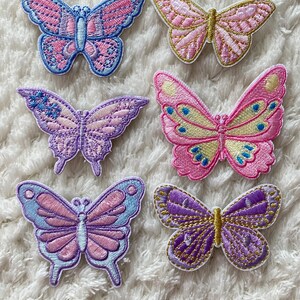 May include: Six embroidered butterfly patches in various colours, including pink, purple, and blue. The patches are on a white fluffy surface.