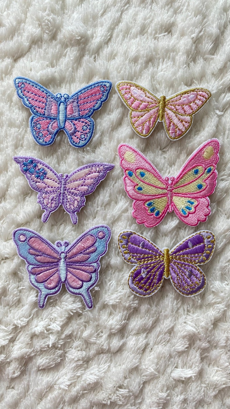 May include: Six embroidered butterfly patches in various colours, including pink, purple, and blue. The patches are on a white fluffy surface.