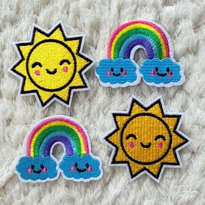 Può includere: Quattro toppe ricamate raffiguranti un sole giallo e un arcobaleno con nuvole blu. Il sole e le nuvole hanno facce sorridenti. Le toppe sono gialle, blu, rosa e arcobaleno con contorni neri.