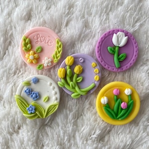 Può includere: Cinque bottoni rotondi in resina colorati con motivi floreali. I bottoni presentano diversi fiori, tra cui tulipani, margherite e un singolo fiore bianco con uno stelo verde. Un bottone ha la parola "Love" scritta su di esso.