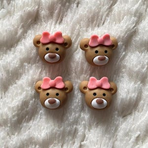 Könnte beinhalten: Vier braune, bärenförmige Knöpfe mit rosa Schleifen. Die Knöpfe sind aus einem weichen, biegsamen Material gefertigt und haben ein süßes, cartoonartiges Aussehen.